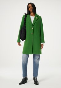Groene wollen jas met gesneden revers en twee knopen. Gecombineerd met lichtblauwe jeans en zwarte enkellaarzen. Zwarte schoudertas met een gewatteerde textuur.
