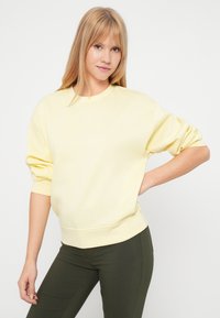 Šviesiai geltona sportinė palaidinė su apvalia iškirpte ir nuleistomis petnešomis, pagaminta iš minkštos medžiagos. Derinama su tamsiai žaliomis prigludusiomis kelnėmis.