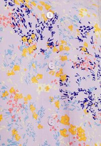 Tissu violet clair avec des motifs floraux jaunes, roses et bleus. Doté de boutons blancs et d'une texture lisse. Manche courte.
