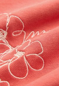 Korallenfarbener Stoff mit weißer floralem Stickerei. Die Textur ist weich mit einem feinen Strickmuster, das die filigranen floralen Details hervorgehoben.