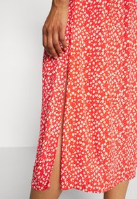 Robe florale rouge en tissu léger, ornée de motifs de fleurs blanches et roses, avec une fente latérale pour faciliter les mouvements.