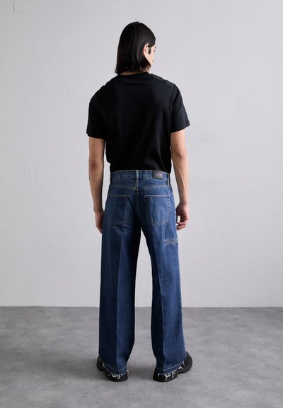 Fekete póló, rövid ujjú, vállrészletekkel, amelyet magas derekú, bőszárú kék farmernadrággal viselnek, hátsó zsebekkel és rojtos szegéllyel.