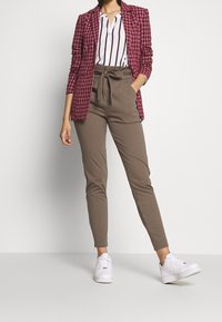 Blazer à carreaux bordeaux porté sur une chemise blanche à rayures, associé à un pantalon kaki fuselé et des baskets blanches. Lignes épurées, silhouette ajustée.