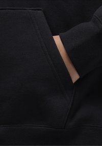 Felpa nera con tasca a marsupio, polsini a costine e una texture liscia. Il materiale appare morbido ed elastico. Una mano è parzialmente visibile.