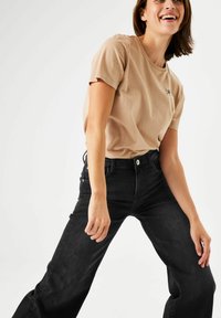 T-shirt en coton beige clair à manches courtes, col rond, avec un petit détail graphique sur la poitrine, associé à un jean en denim noir.