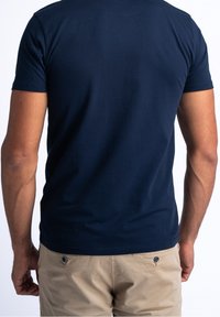 Tmavě modré bavlněné tričko s krátkými rukávy, přiléhavým střihem a hladkou texturou. Nošeno s lehce béžovými šortkami s knoflíkovým detailem.