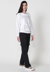 Felpa bianca con testo in paillettes argento "BRAVE & FREE," abbinata a pantaloni cargo neri con tasche a toppa e dettagli in argento.