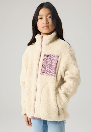 Jong persoon draagt een crèmekleurig fleecejack met een roze luipaardprint borstzak en rits, handen in de zakken, staand tegen een neutrale achtergrond.
