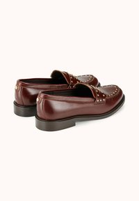 Mocassins en cuir marron avec accents cloutés, bout arrondi, texture lisse et petit talon empilé. La couture décorative met en valeur le design.