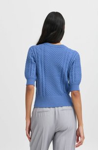 Hellblau gestrickter Pullover mit strukturierten Kabel- und Wabenmuster, kurzen Puffärmeln und geripptem Saum, kombiniert mit grauen Hosen.
