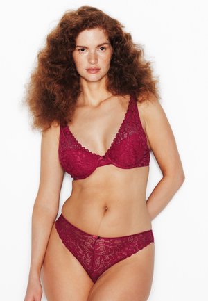 Donna con capelli rossi ricci che indossa un reggiseno in pizzo rosso e mutandine abbinate, in piedi davanti a uno sfondo bianco con espressione neutra.