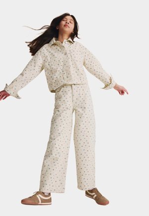 Young woman with long hair wearing cream floral jacket and pants, standing with arms slightly extended and eyes closed.
