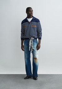 Homme portant une chemise à manches longues rayée bleu marine avec le texte « Rebels », un jean patchwork, des chaussures noires et une écharpe à motif nouée autour de la taille.