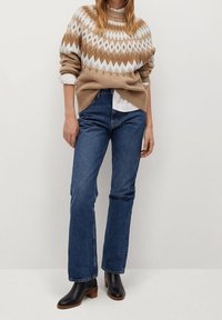 Beige und weiße gemusterte Strickjacke über einem weißen Hemd, kombiniert mit blauen geraden Jeans und schwarzen Lederknöchelstiefeln.