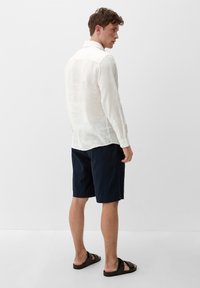 Weißes Leinenhemd mit langen Ärmeln, getragen mit navyfarbenen Shorts und schwarzen Sandalen. Das Hemd hat einen klassischen Kragen und einen strukturierten, leichten Stoff.