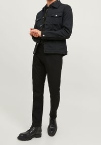 Chaqueta de denim negra con dos bolsillos frontales, herrajes plateados y cierre de botón. Combinada con pantalones negros y botas negras de cordones.