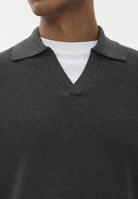 Sötétszürke kötött pulóver V-nyakkal és rövid gallérral. A textúrája bordázott; egy fehér póló fölé van rétegezve.