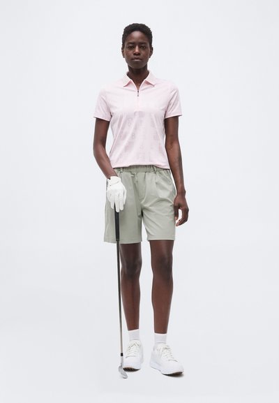 Femme portant un polo rose clair et un short vert clair, tenant un club de golf avec un gant blanc sur un fond uni.