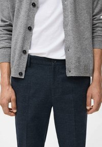 Grijze gebreide cardigan met zwarte knopen over een witte T-shirt, gecombineerd met donkerblauwe gestructureerde broeken met een randknopsluiting.