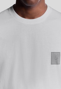 Hvid bomulds T-shirt med rund halsudskæring og et lille gråt logo-patch med en due og "1874" i nederste højre hjørne.