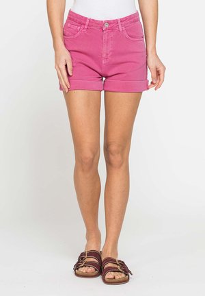 Donna con pantaloncini di jeans rosa con orlo arrotolato e sandali marroni, in piedi su uno sfondo chiaro e uniforme.