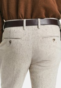 Nahaufnahme von beiger gepunkteter Hose mit geknöpften Gesäßtaschen, getragen mit einem dunkelbraunen Ledergürtel und einem braunen Pullover, der in die Hose gesteckt ist.