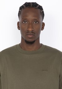 T-shirt à manches longues vert olive en coton doux. Présente un col rond et un petit logo discret sur la poitrine.