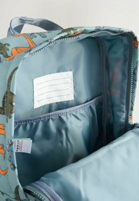 Ryggsäck med blådinosaurmotiv, zippered huvudfack, invändig ficka och namnlapp. Mjuk tyg och hållbar sömnad är synlig.