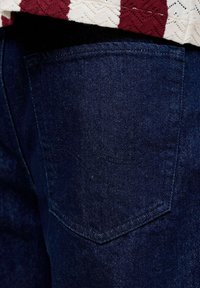 Πίσω τσέπη σκούρου μπλε τζιν με ορατό ράψιμο, σε συνδυασμό με λευκή και κόκκινη πλεκτή άκρη πουλόβερ.