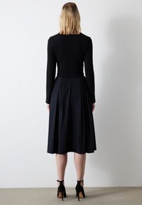 Schwarzes langärmliges Kleid mit eng anliegendem Oberteil und plissiertem Rock. Der Stoff sieht glatt aus und hat ein stylisches Rückendetail. Schwarze Absätze mit Knöchelriemen.