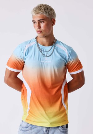 Jonge man met kort blond haar die een sportshirt draagt met een blauwe naar gele kleurverloop, zilveren kettingen en lichte spijkerbroek, handen achter de rug.