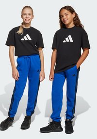 T-shirts noirs avec le logo Adidas blanc, pantalons de survêtement bleus avec des rayures latérales noires, et chaussures de sport noires. Matériau en coton, coupe décontractée.