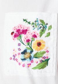 Design floral brodé mettant en vedette des fleurs roses, jaunes et bleues sur un fond en tissu blanc, avec des accents en perles et des feuilles vertes.