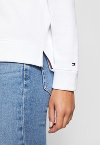 Gros plan d'une personne portant un jean bleu clair et une chemise blanche à manches longues avec un petit logo sur le poignet et une fente latérale.