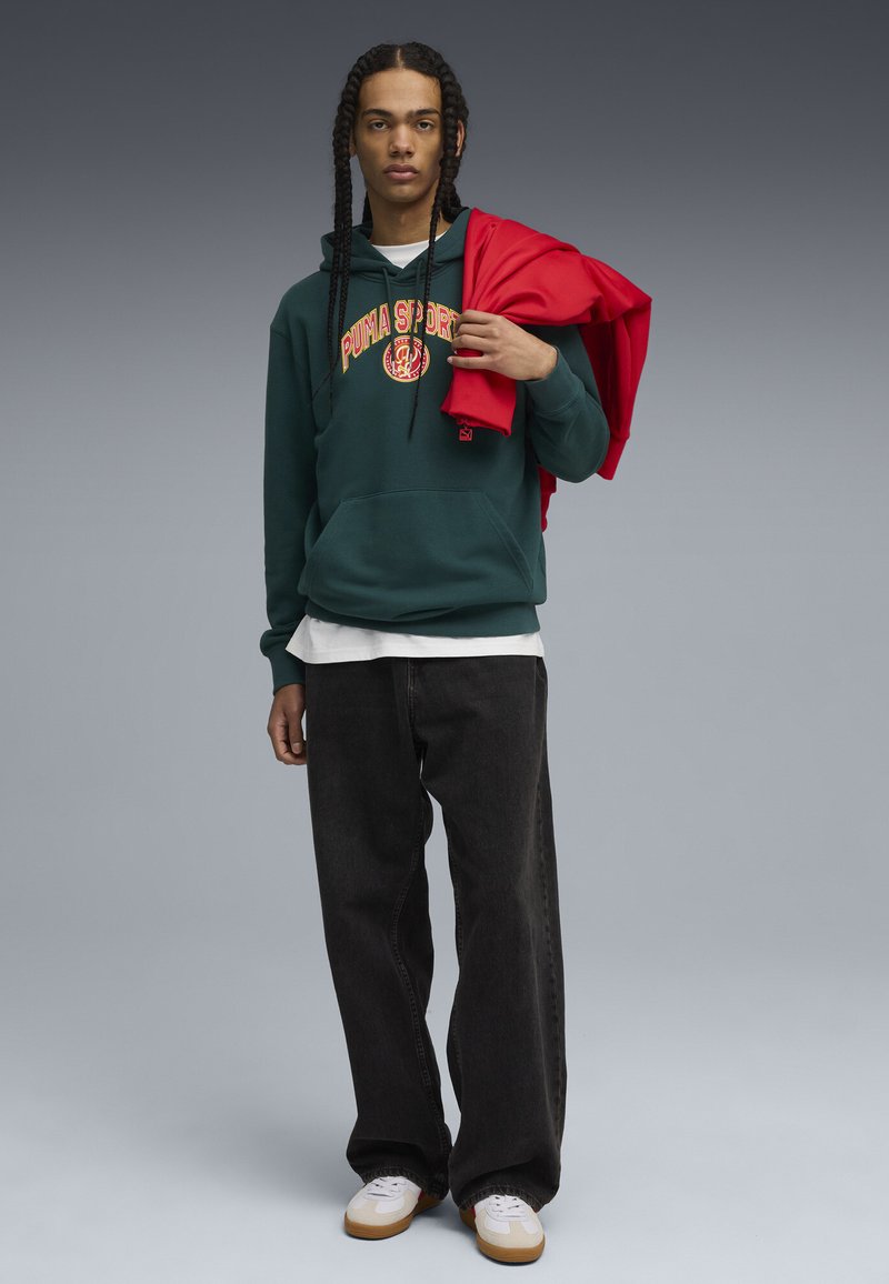 Sudadera verde oscuro con logo rojo, camiseta blanca, jeans negros y zapatillas blancas. Modelo sosteniendo una chaqueta roja sobre su hombro.