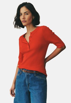 Ribbad, kortärmad röd topp med knäppning i halsringningen, i kombination med blå denimjeans och ett brunt bälte. Tygerna ger en texturerad look.