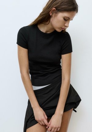 Fiatal nő fekete rövid ujjú felsőben és fekete szoknyában, lehajtott fejjel, előtte összekulcsolt kézzel, egyszerű háttér előtt.