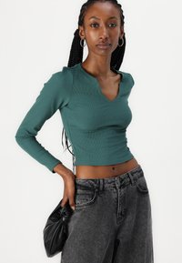 Teal ribbet langermet topp med V-hals, kombinert med høy midje mørkegrå jeans. Modellen holder en svart veske med knyttet design.