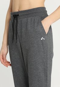 Pantalones de chándal grises con cintura elástica y cordón. Cuenta con bolsillos laterales y un pequeño logo blanco en el muslo. Textura de tela suave.