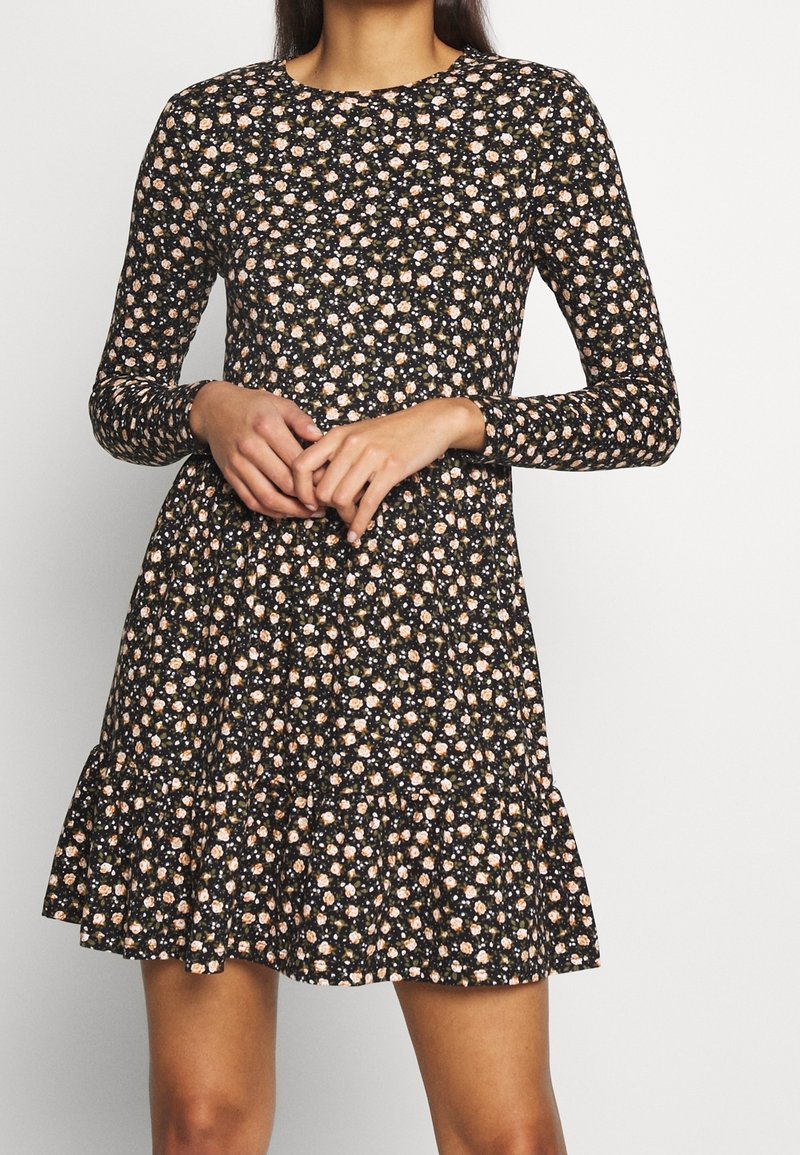 Robe noire à manches longues avec un motif floral rose et blanc, dotée d'un col rond et d'une jupe évasée avec un ourlet volanté.