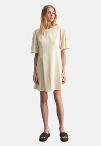 Hellgelbes, kurzärmliges Kleid mit Rundhalsausschnitt und ausgestelltem Saum, gefertigt aus einem glatten Stoff. Getragen mit dunklen Schlüpfschuhen.