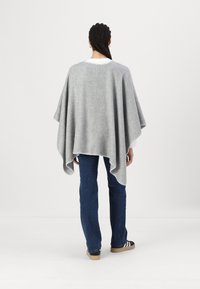 Poncho de lã cinza com um formato solto, bordas com bainha e detalhes de costura. Usado sobre uma camisa branca e combinado com jeans azuis e ténis.