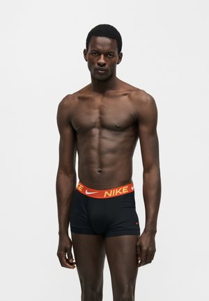 Atletický tmavší muž stojaci, oblečený v čiernych boxerkách Nike s oranžovým pásom a logom na jednofarebnom svetlom pozadí.