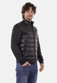 Homme aux cheveux bouclés portant une doudoune noire sans manches et un t-shirt noir à manches longues, debout avec une main dans la poche devant un fond uni.