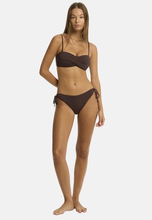 Młoda kobieta stoi boso, ubrana w ciemnobrązowe bikini z wiązanymi z boku majtkami na jednolitym białym tle.