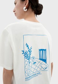 Witte katoenen t-shirt met een blauwe grafische print van een tafel met een vaas, bladeren en een stoel, met de tekst "mediterranean soul" onderaan.