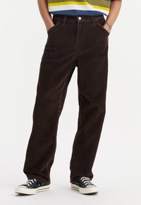 Pantalones de pana marrón de corte recto, con bolsillos laterales y cierre de botón frontal. Combinados con zapatillas negras de caña baja.