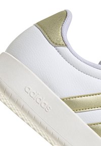 Zapatilla blanca de cuero con acentos dorados, panelado texturizado y suela de goma. Presenta el nombre de la marca bordado en el lateral.