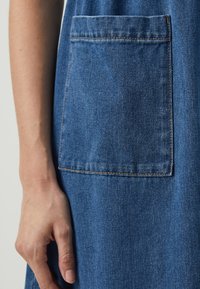Denimkjole med en stor lomme, blått stoff, subtil tekstur og kontraststing langs kantene. Hånd hviler ved siden av lommen.