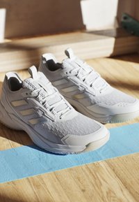 Graue Sportschuhe aus strukturiertem Mesh und synthetischen Materialien. Zu den Merkmalen gehören Schnürsenkel, eine verstärkte Zehenkappe und drei weiße Streifen.